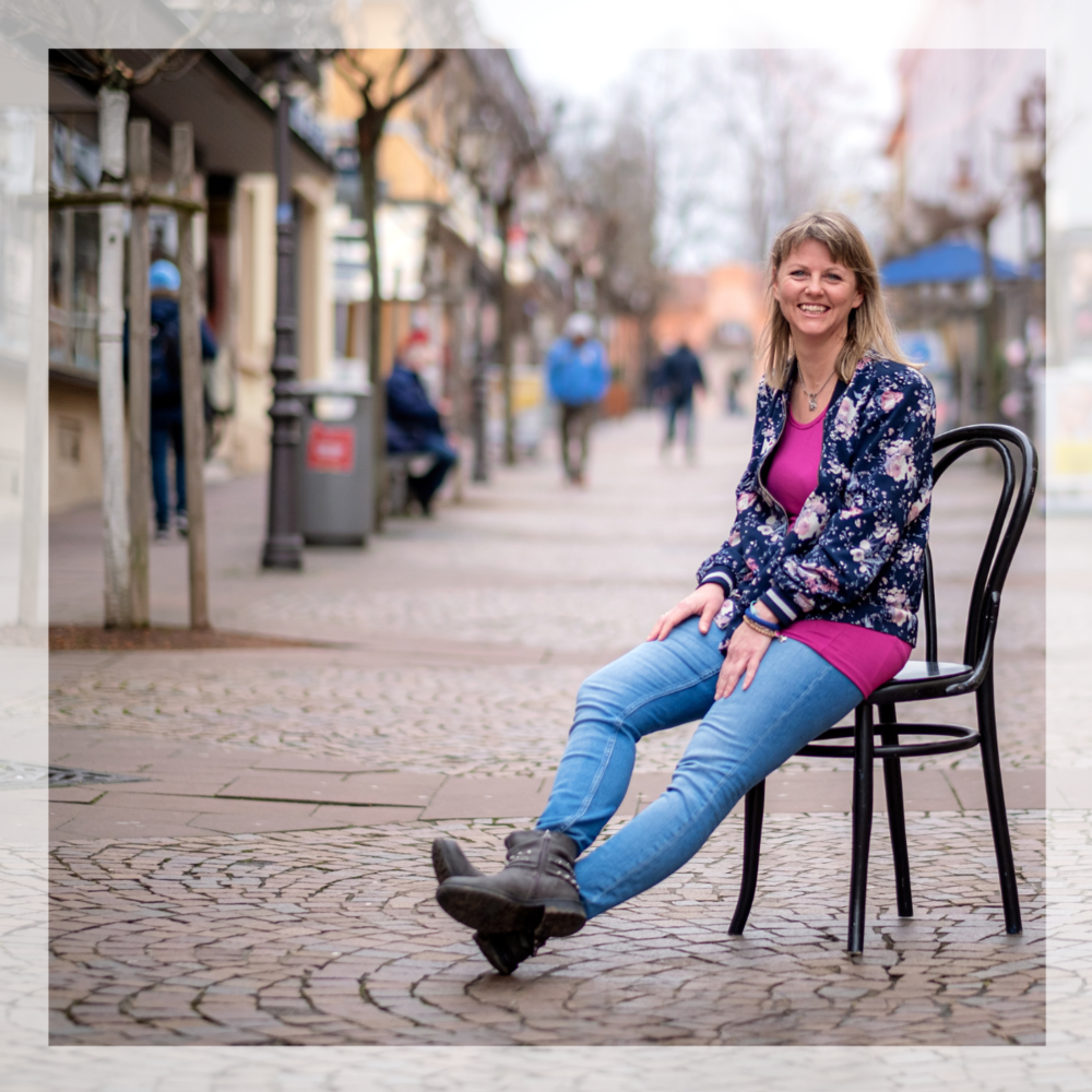 Stress Coach sitzt lachend auf einem Stuhl in der Fußgängerzone